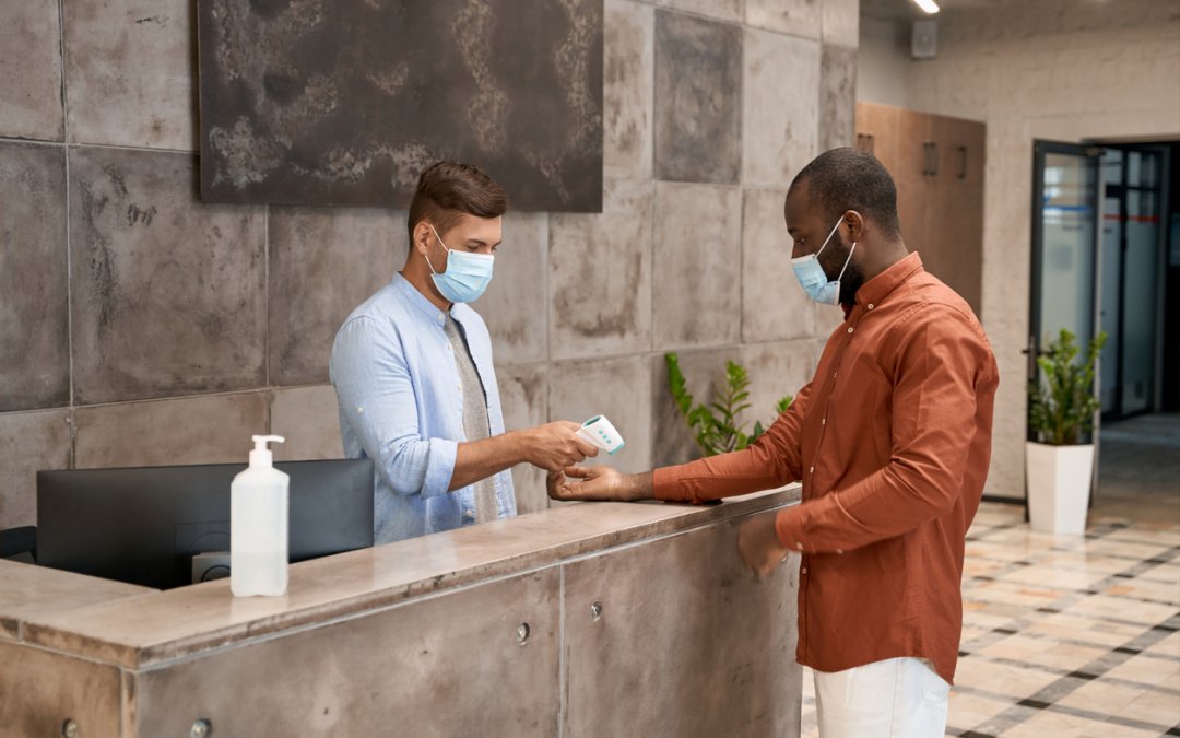Young male receptionist wearing medical mask standing at reception in the modern office and measuring body temperature of man with infrared thermometer